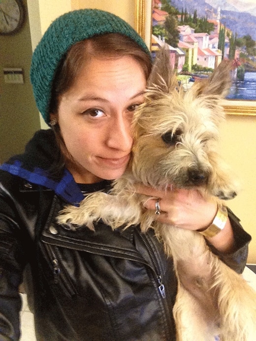 Woman in black leather jacket and green beanie holding her brown terrier.